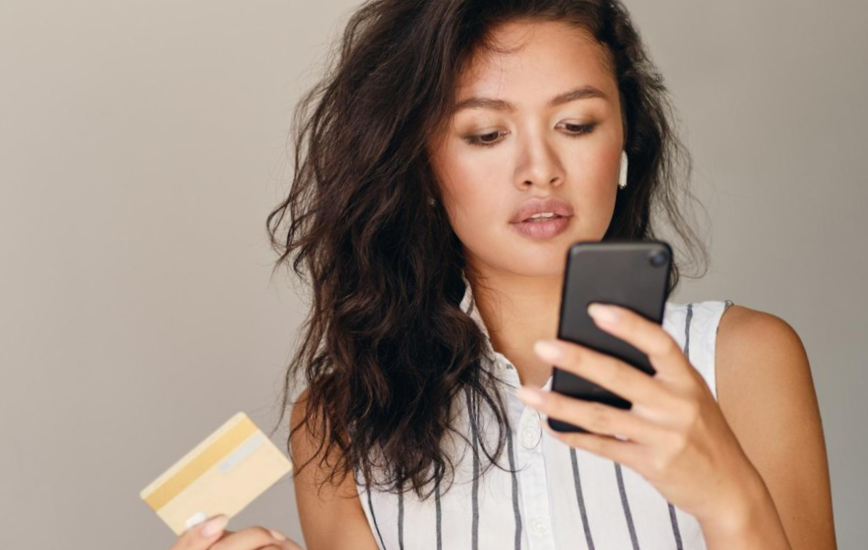 Portrait of young beautiful asian woman with credit card confidently using cellphone over gray background